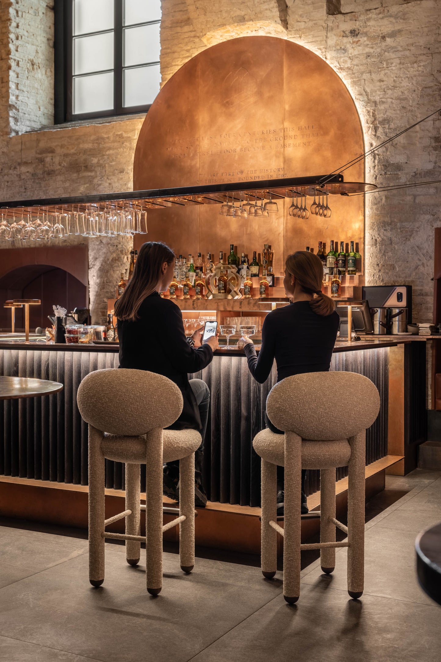 Two women enjoying drinks on modern Flock CS1 bar stools at a stylish bar with warm lighting.