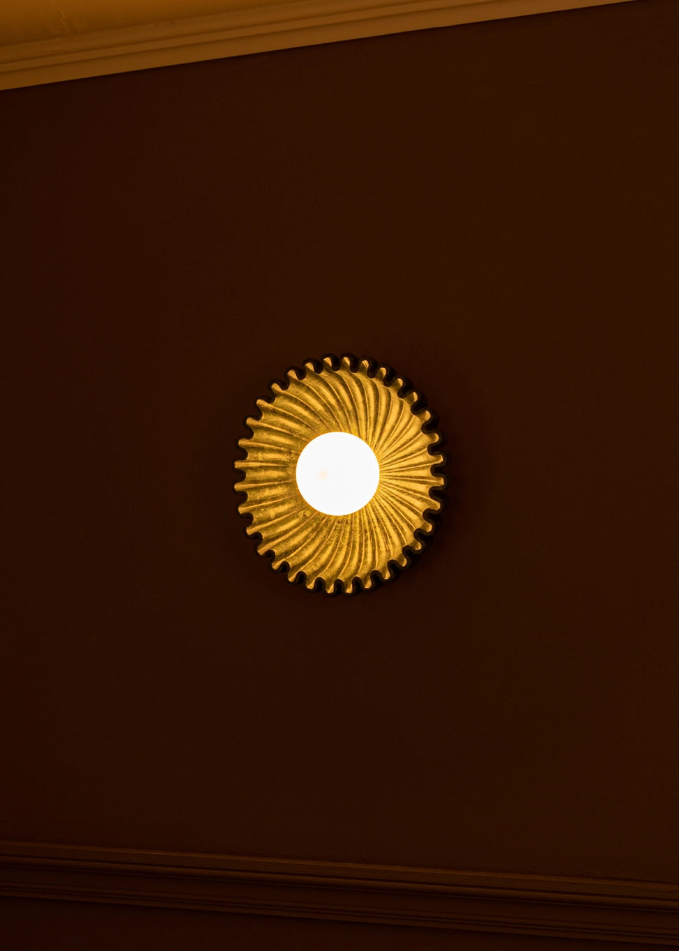 Designer circular wall sconce with sunburst design, glowing softly against a dark wall, adding elegance to any interior.